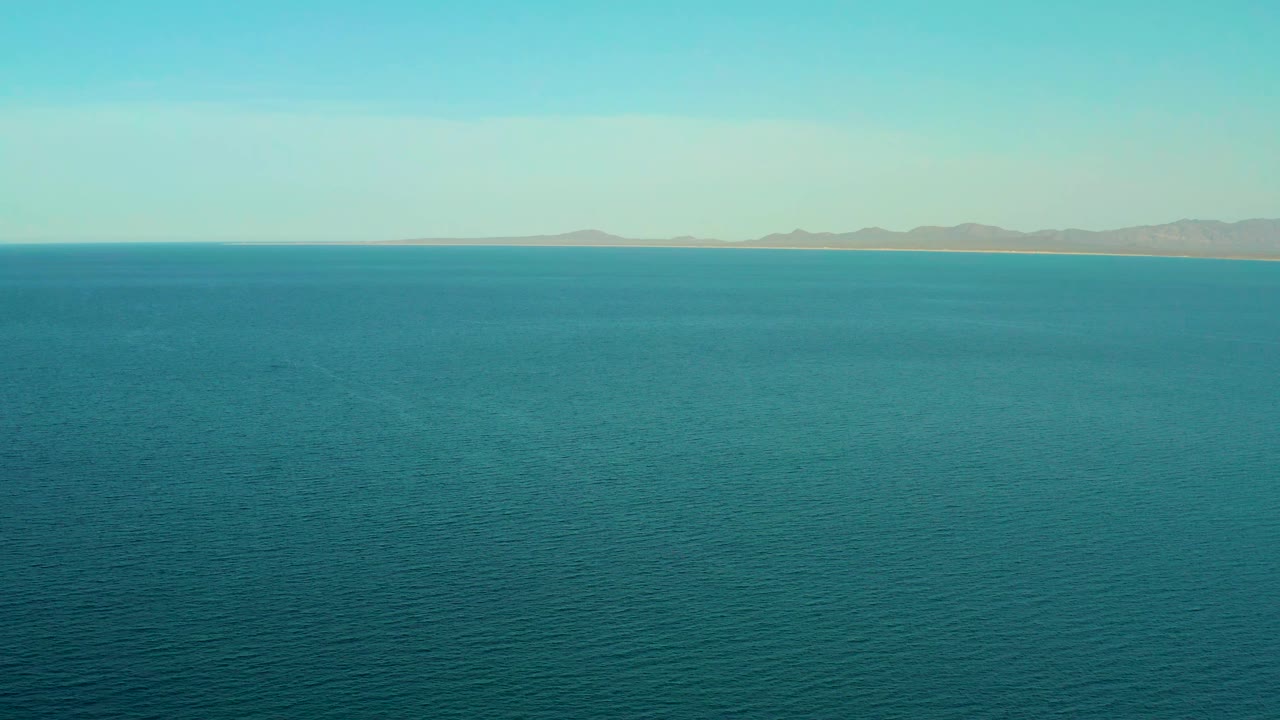 Blue ocean surface fills the foreground and midground, with El Sargento La Ventana's distant coast and low mountain shapes under soft daylight, no visible people.