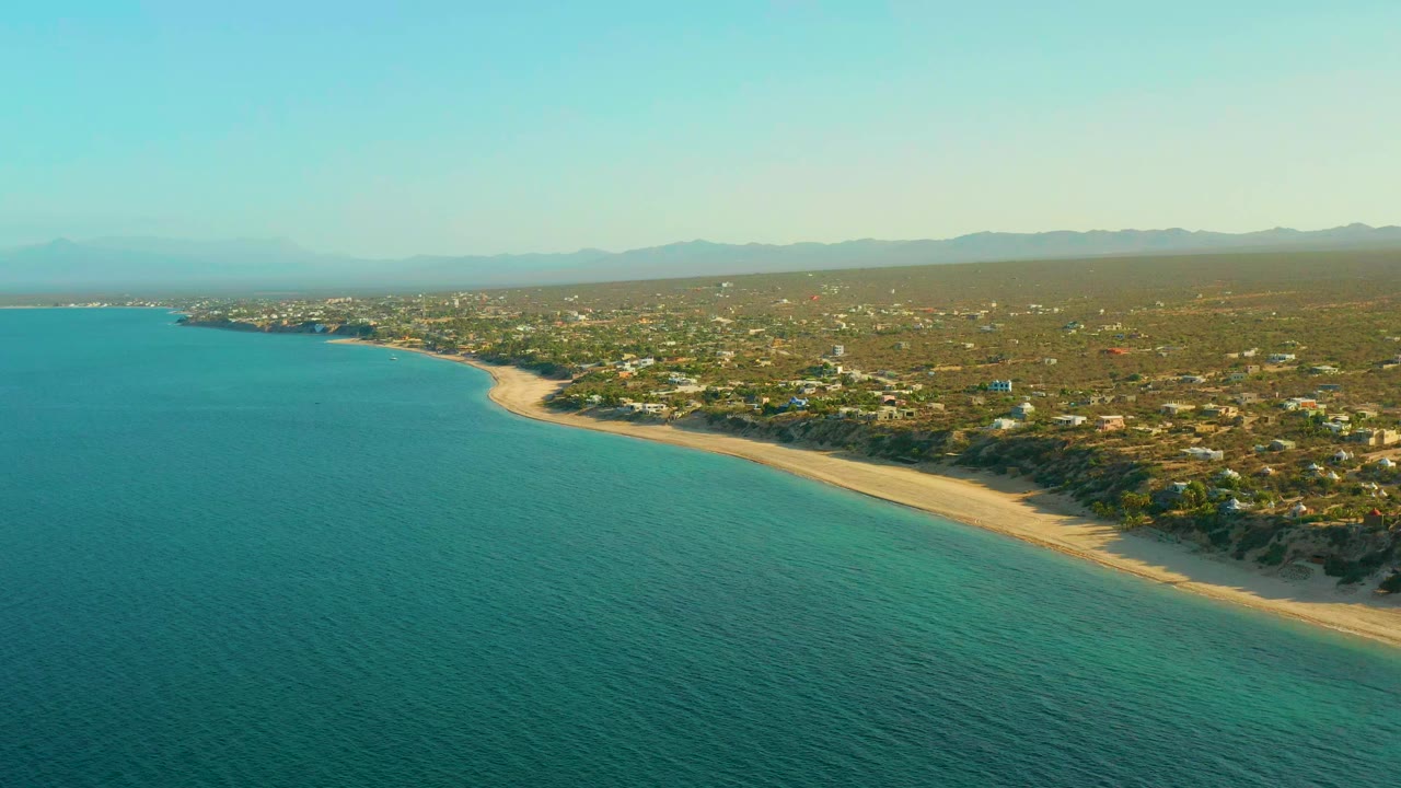Wide aerial view over El Sargento La Ventana, Mexico, with turquoise ocean in the foreground, a curving sand beach, scattered inland homes, distant mountains, and no visible people.