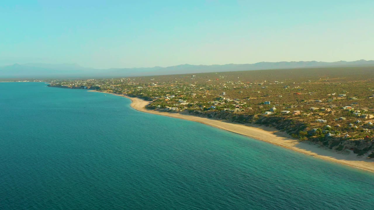 High coastal view with blue-green water, beach curve, houses, and hazy mountains.