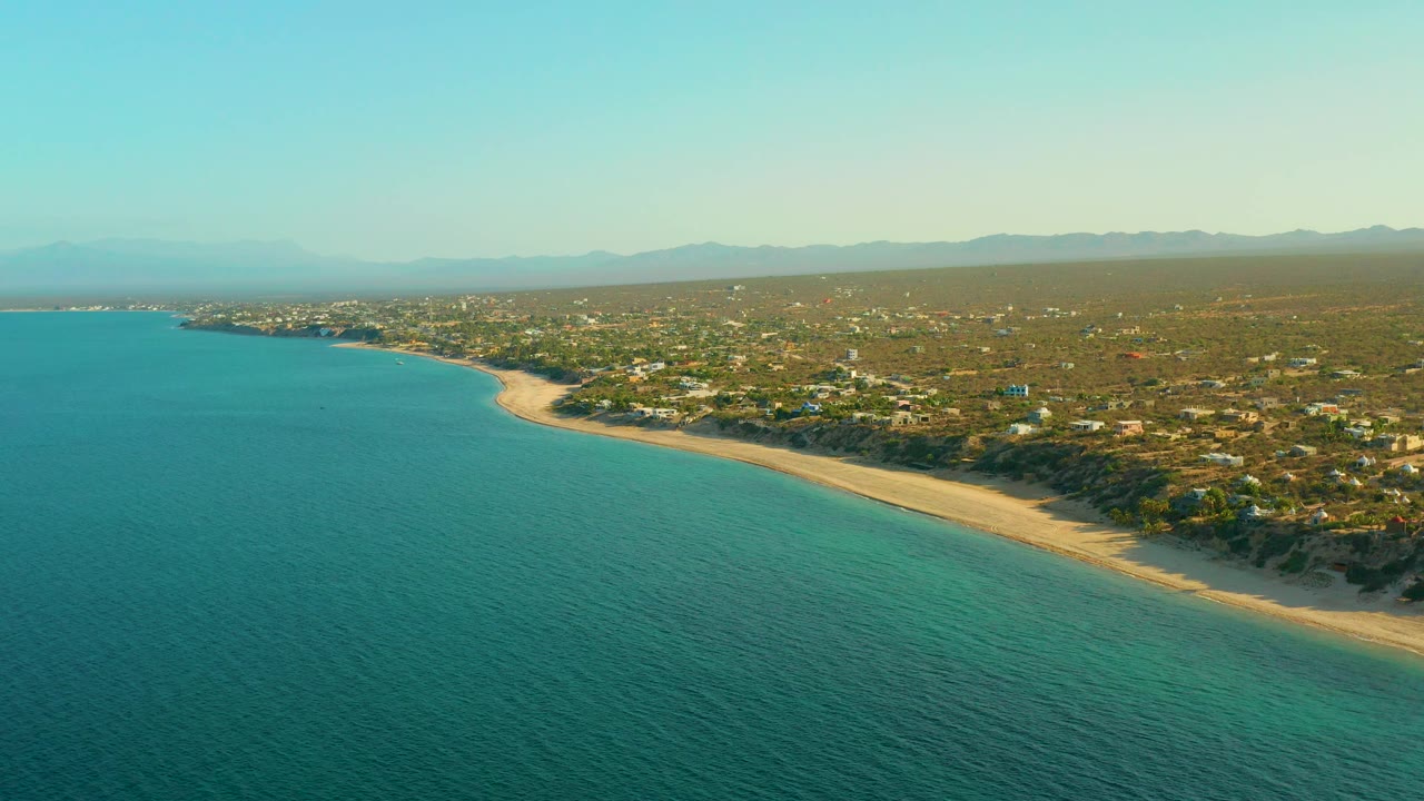 Aerial frame over El Sargento La Ventana, Mexico, with open turquoise water on the left, a long pale beach on the right, inland buildings, distant ridgelines, and no visible people.