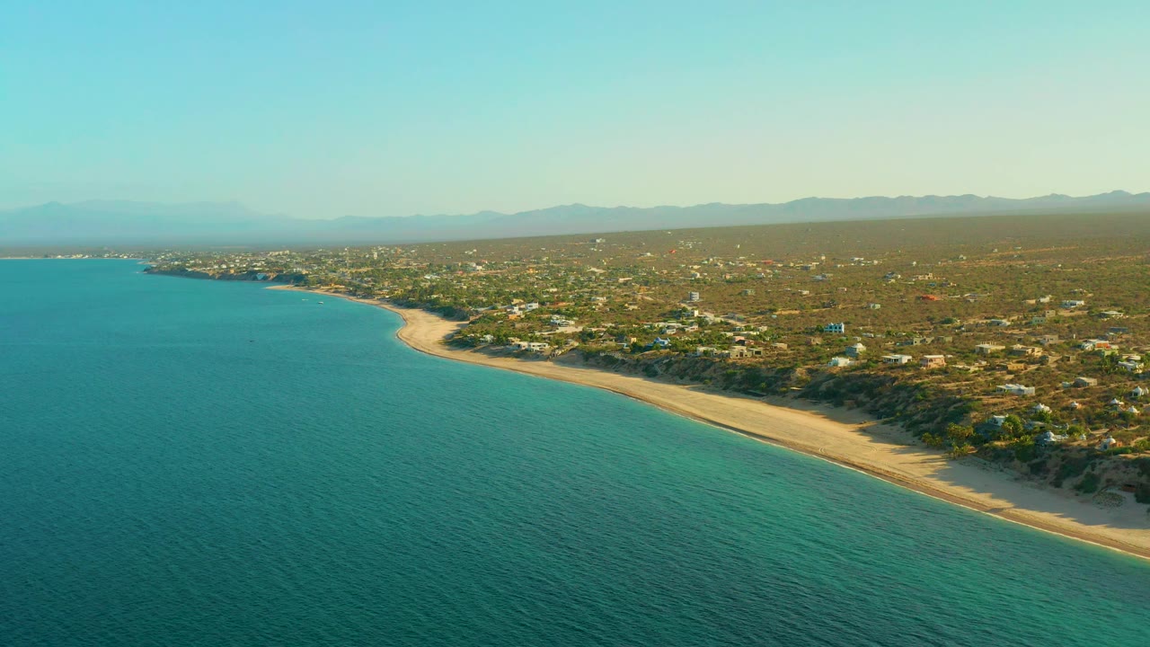 The second frame keeps the El Sargento La Ventana shoreline centered, with turquoise water below, pale sand at the edge, inland homes and roads, distant mountains, and no visible people.