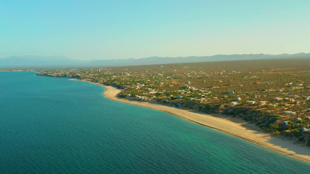 High aerial frame of El Sargento La Ventana, Mexico, with blue-green ocean, a pale beach curve, inland scrub, small structures, and a hazy mountain horizon with no visible people.
