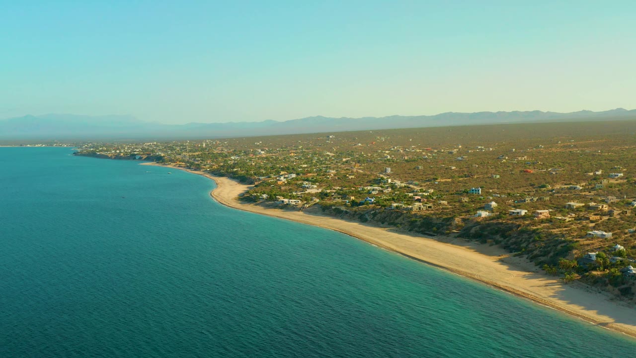 Wide drone view over El Sargento La Ventana, Mexico, where open turquoise water meets a long sandy shoreline, with scrubby inland terrain and distant mountains under soft haze, no visible people.