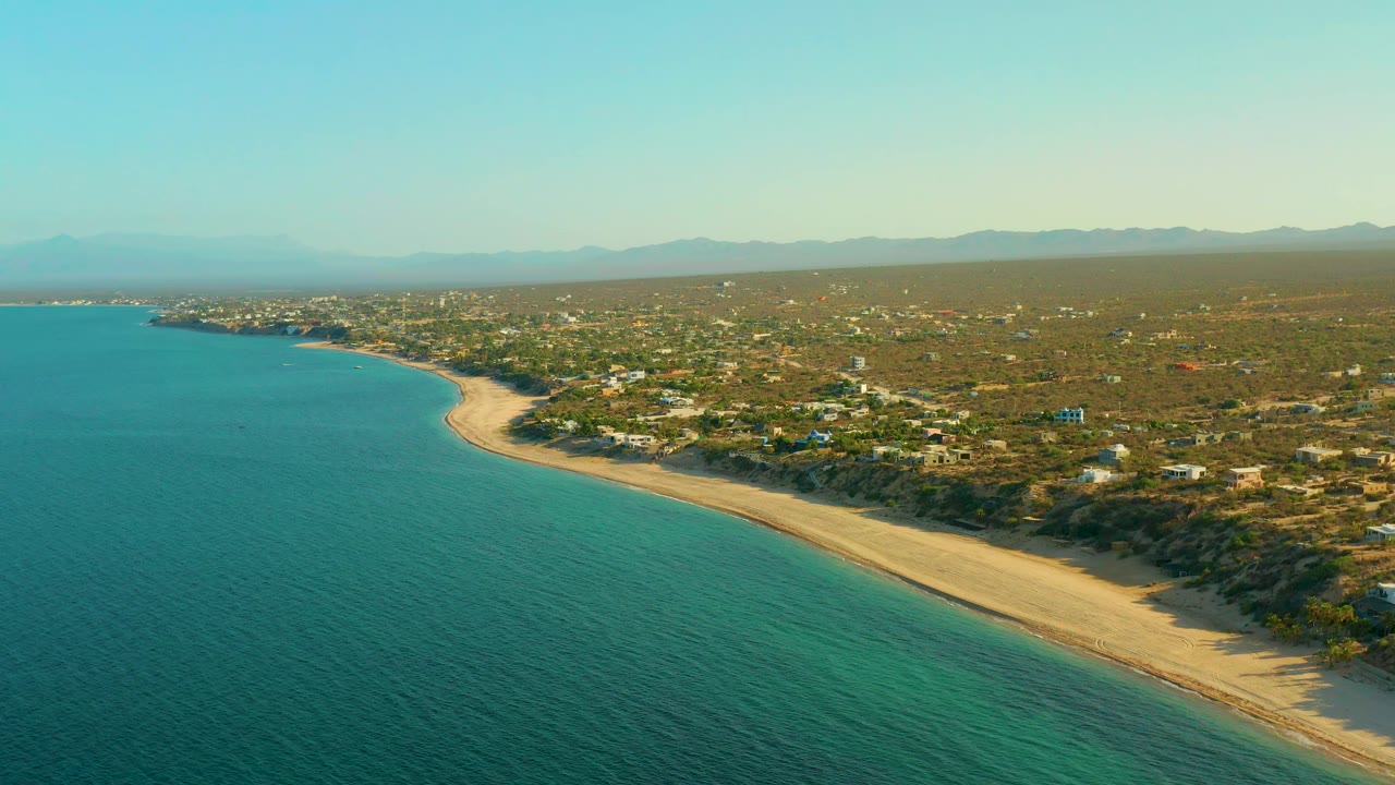 Aerial frame from El Sargento La Ventana, Mexico, showing blue-green water along the left side, a long sand beach curving inland, low buildings across scrubland, distant mountains, and no visible people.