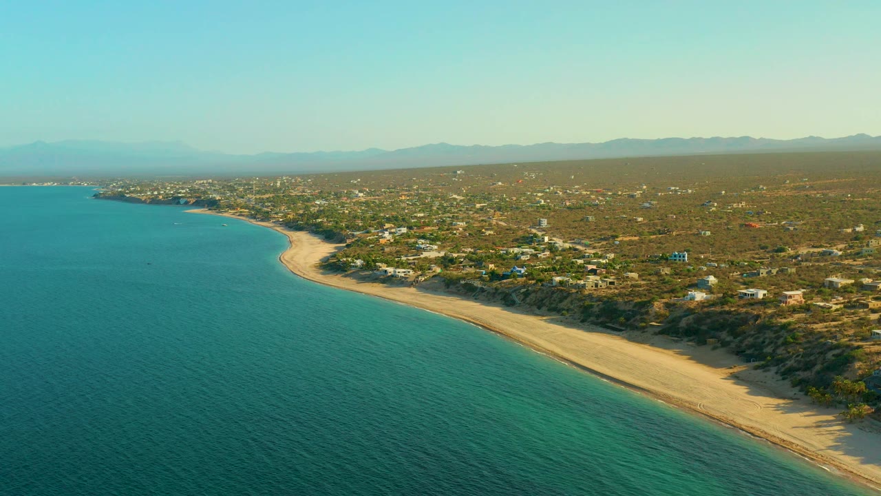 High aerial view over El Sargento La Ventana, Mexico, with shallow turquoise water in the foreground, a pale beach bending around the bay, coastal houses in scrub terrain, distant mountains, and no visible people.