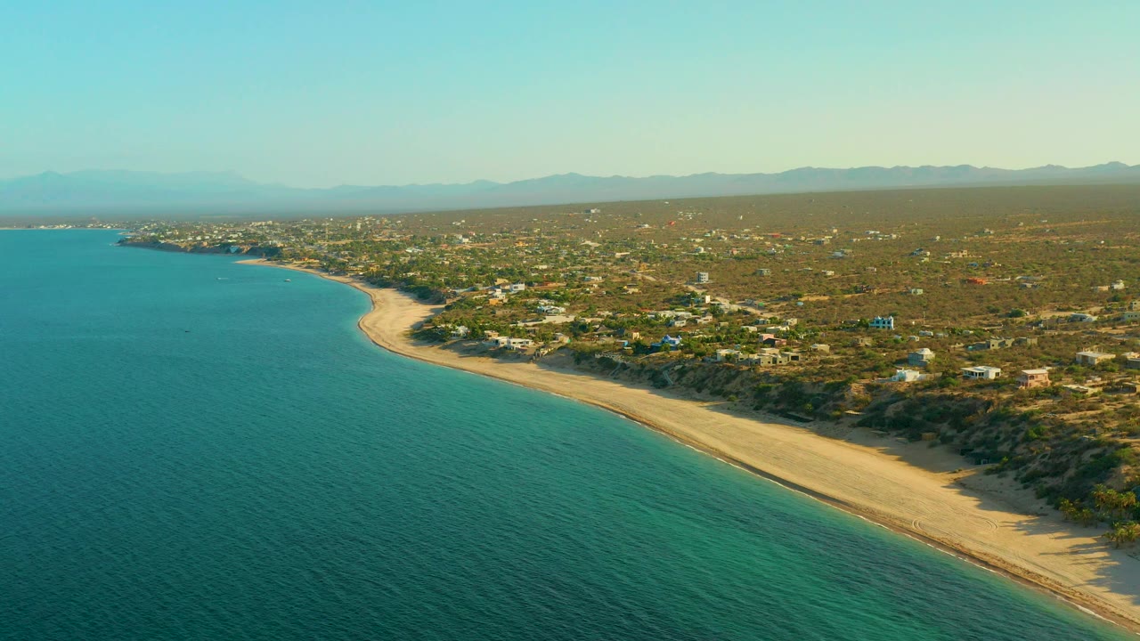 Drone view of curved beach beside calm ocean with homes and dry terrain inland.