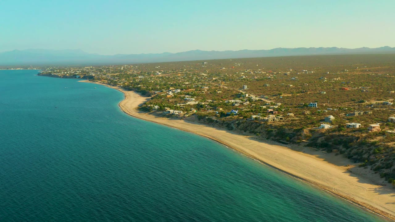 High coastal view with blue-green water, pale sand, dry terrain, and small buildings inland.