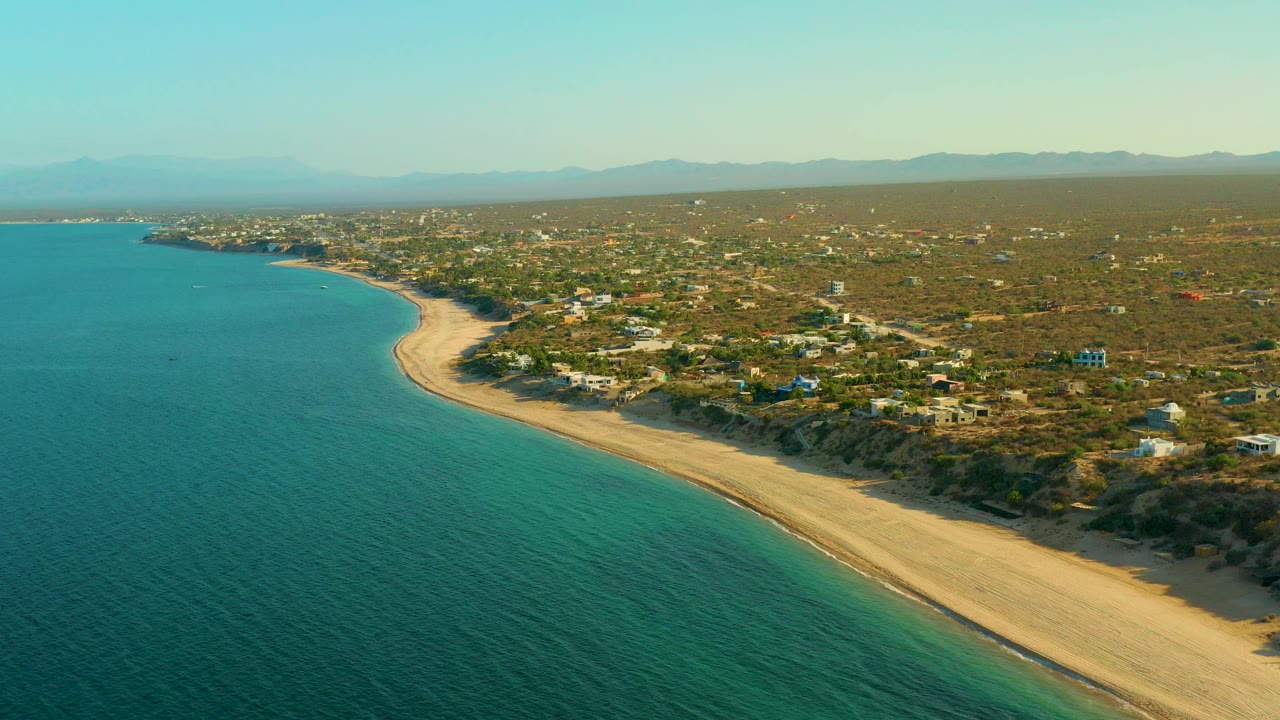 Opening aerial frame over El Sargento La Ventana, Mexico, with turquoise water on the left, a pale curved beach on the right, scattered buildings inland, hazy mountains on the horizon, and no visible people.