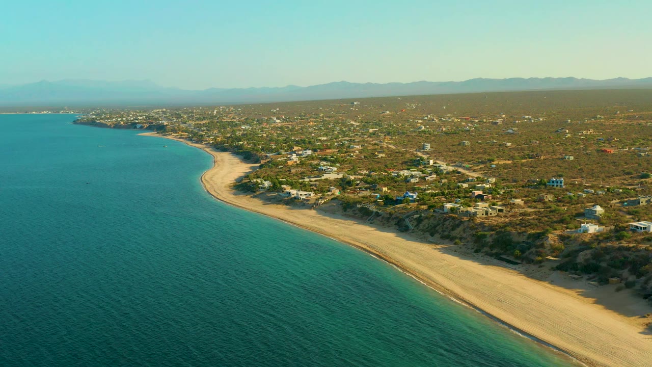 High coastal view with blue-green water, a long beach curve, inland buildings, scrubland, and mountains.