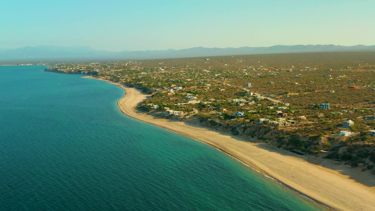 Aerial view of El Sargento La Ventana, Mexico, with calm textured water at left, an empty-looking sand beach at lower right, scattered houses and roads inland, hazy mountains, and no visible people.