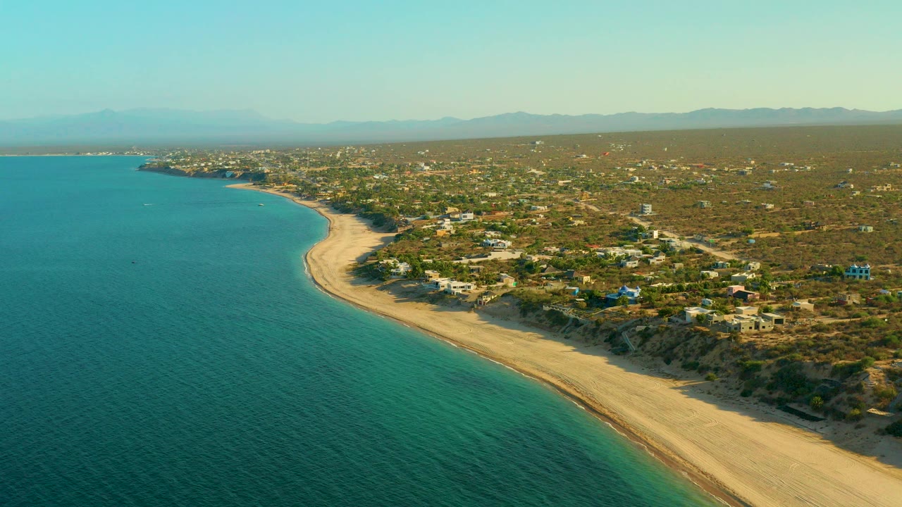 A high aerial frame over El Sargento La Ventana, Mexico, shows turquoise water on the left, a pale sandy shoreline curving through the center, scattered homes inland, hazy mountains in the distance, and no visible people.