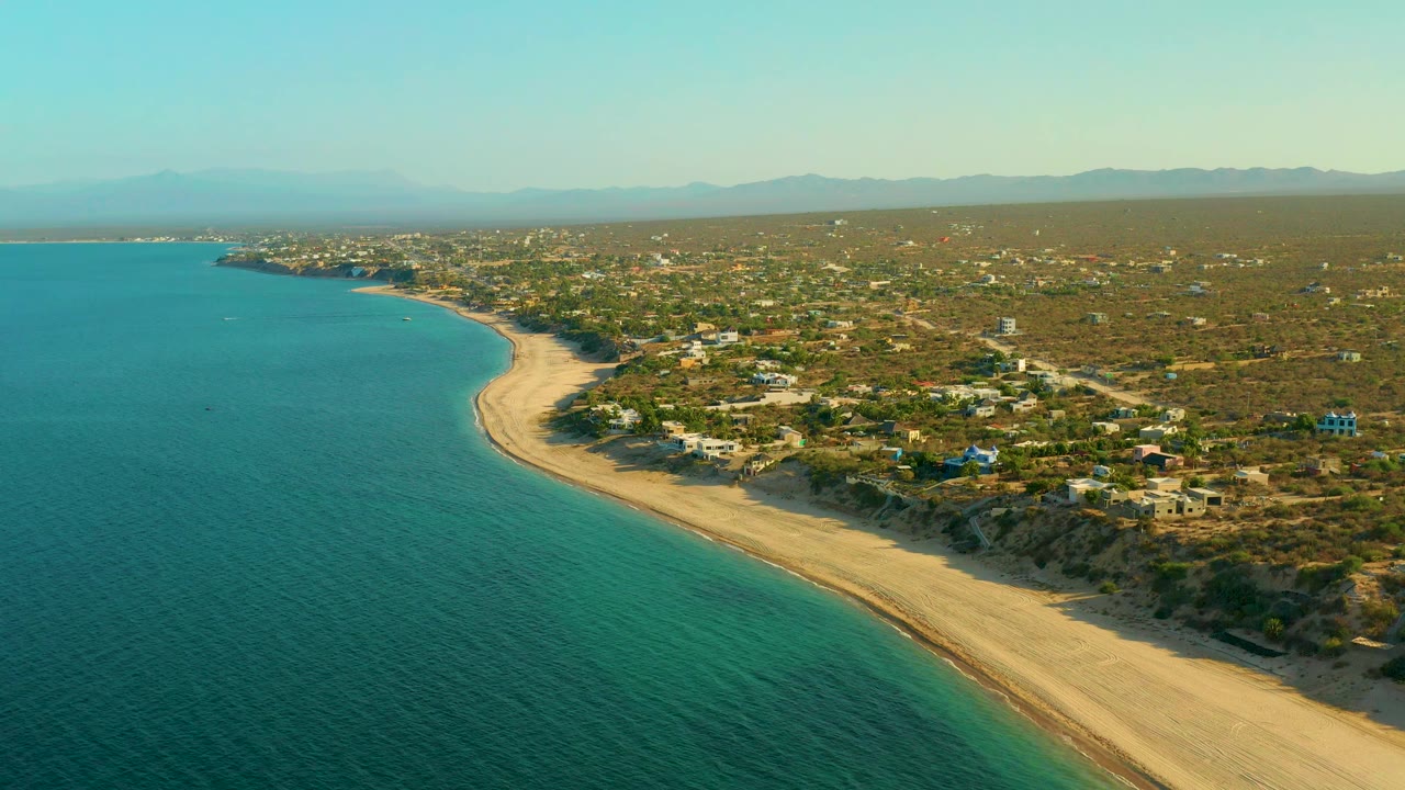 At 0:01, the El Sargento La Ventana shoreline remains in a high aerial composition, with blue-green sea in the foreground, a broad pale beach, low vegetation and houses inland, distant mountains, and no visible people.