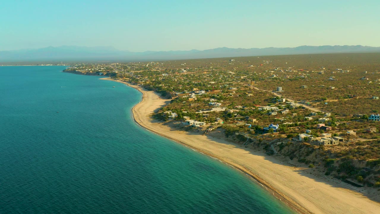 Drone-height view of calm water, a sweeping beach, scattered houses, and desert vegetation.