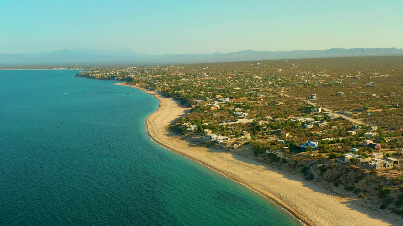 High aerial view over El Sargento La Ventana, Mexico, with turquoise water on the left, a curving sand beach, scattered homes in low vegetation, hazy mountains, and no visible people.