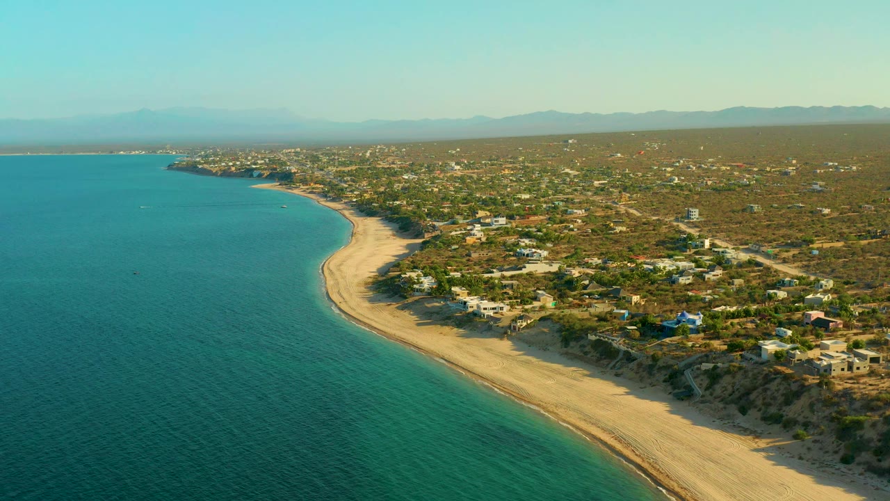 Aerial frame over El Sargento La Ventana, Mexico, looking along the curved beach with open blue-green water, homes set back from dunes, tree-covered terrain, distant mountains, and no visible people.