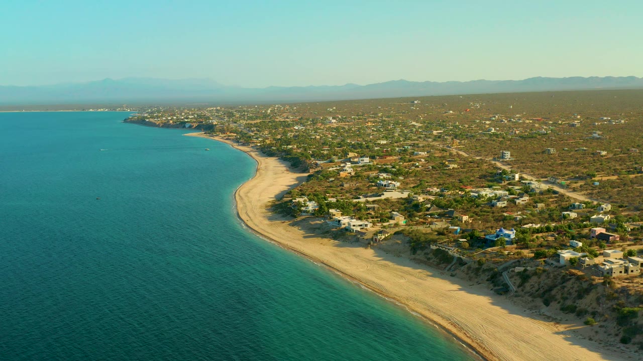 High drone view of El Sargento La Ventana, Mexico, with calm water occupying the left frame, a pale beach running into the distance, homes and roads across dry vegetation, mountains on the horizon, and no visible people.