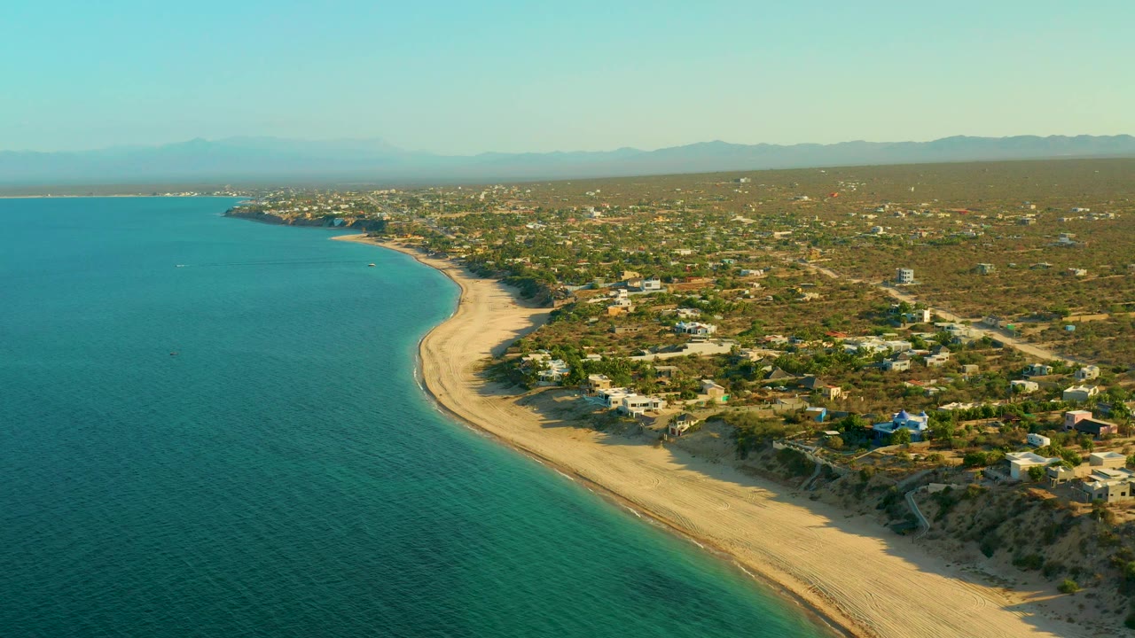 Aerial shoreline view with beach dunes, ocean, houses, vegetation, and mountain backdrop.