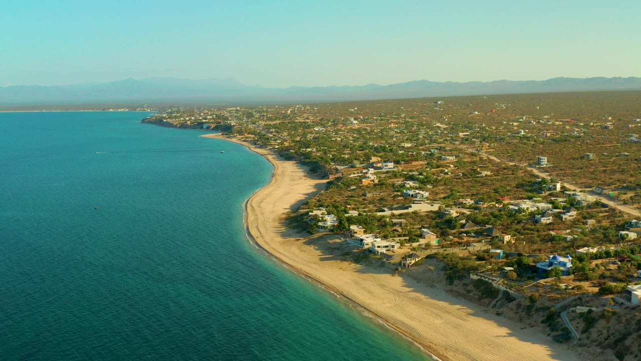 Opening aerial view over El Sargento La Ventana, Mexico, with aquamarine water on the left, a curving sandy beach, scattered inland homes, hazy mountains, and no visible people.