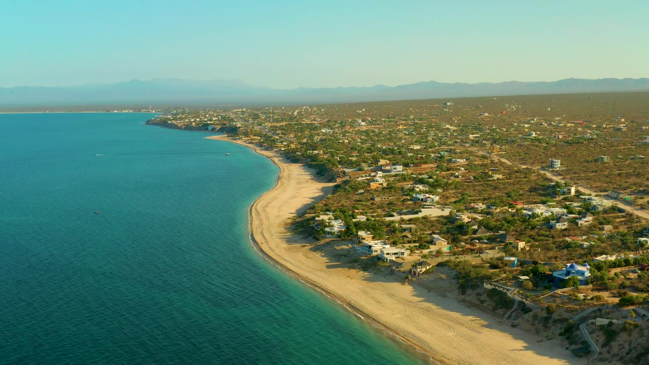 A high aerial frame of El Sargento La Ventana, Mexico, showing teal ocean, a pale beach running diagonally, clustered small homes to the right, dry scrubland, distant mountains, and no visible people.