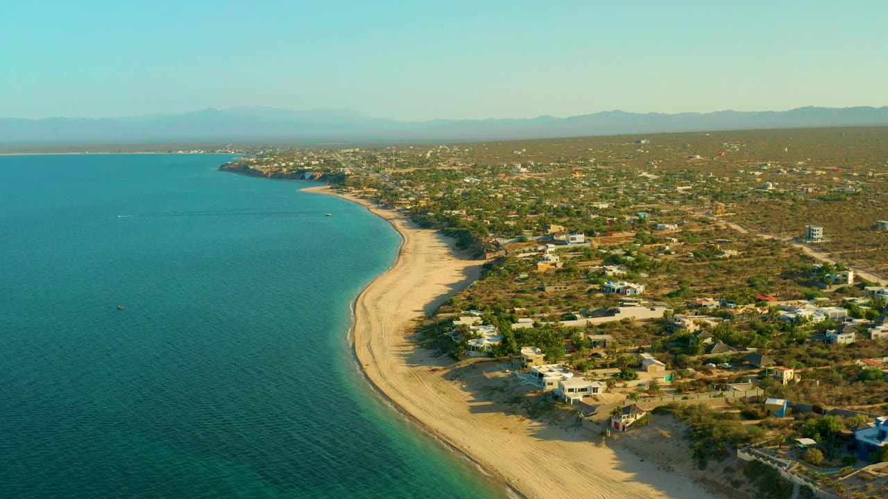 High aerial frame of El Sargento La Ventana, Mexico, showing turquoise water, pale sand, shoreline trees, scattered low buildings, hazy mountains, clear sky, and no visible people.