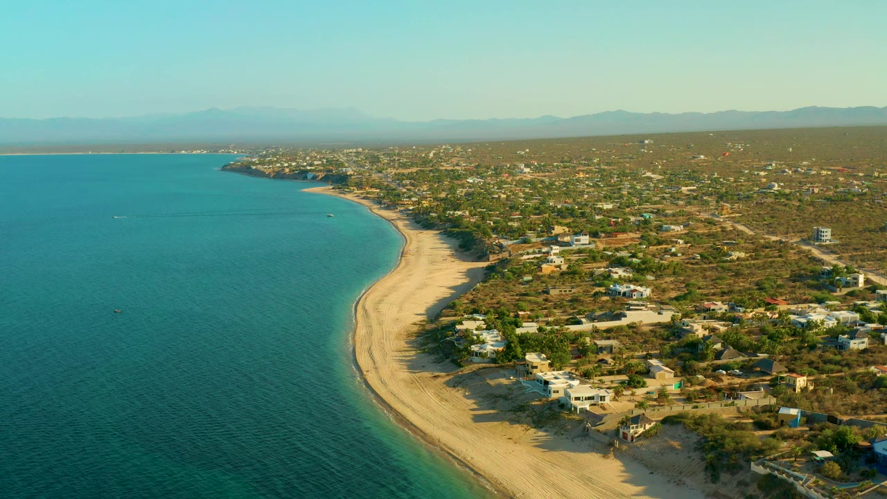 Drone-like aerial view over El Sargento La Ventana, Mexico, with the blue sea filling the left side, a sandy shoreline sweeping through the frame, coastal houses, trees, mountains, and no visible people.