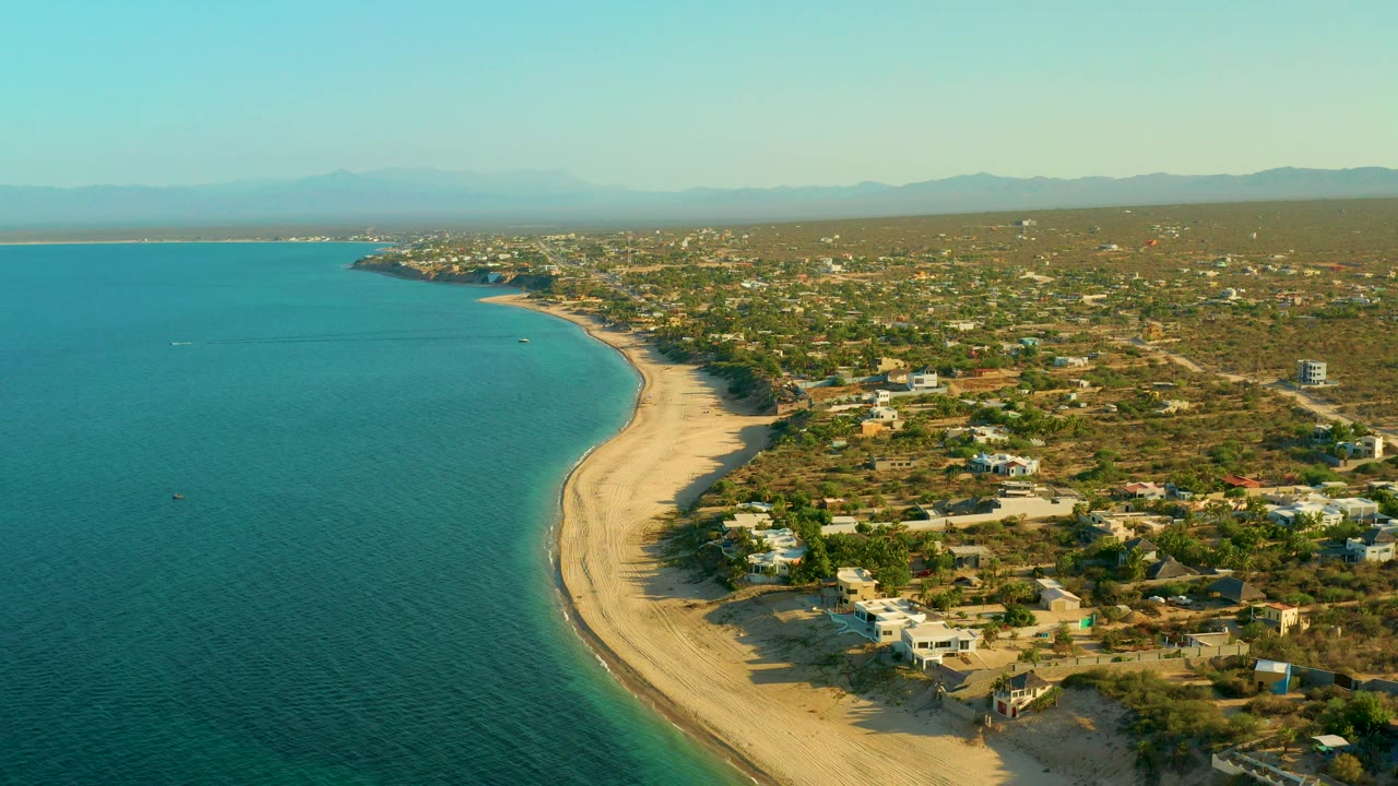 Wide aerial coastline with calm water, beach, trees, homes, and mountains.