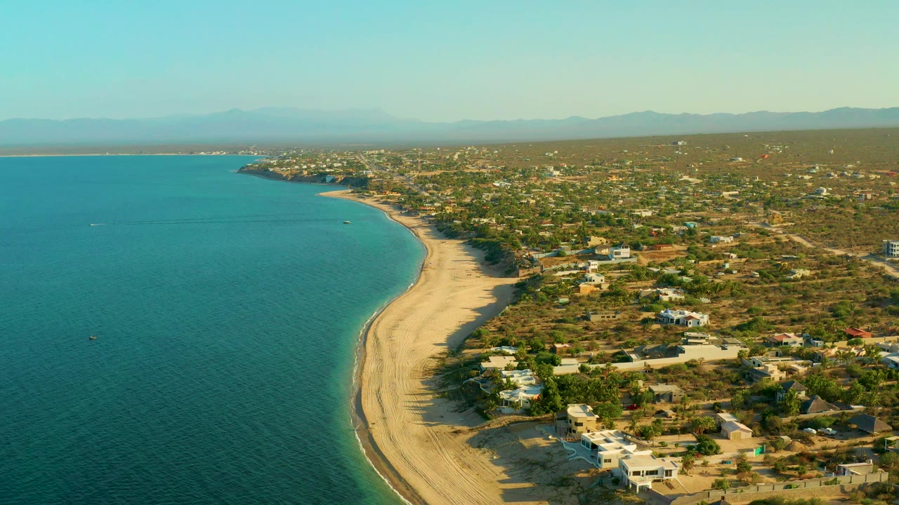 Aerial frame over El Sargento La Ventana, Mexico, looking along the beach as turquoise water meets pale sand, with low homes and scrubland inland and no visible people.
