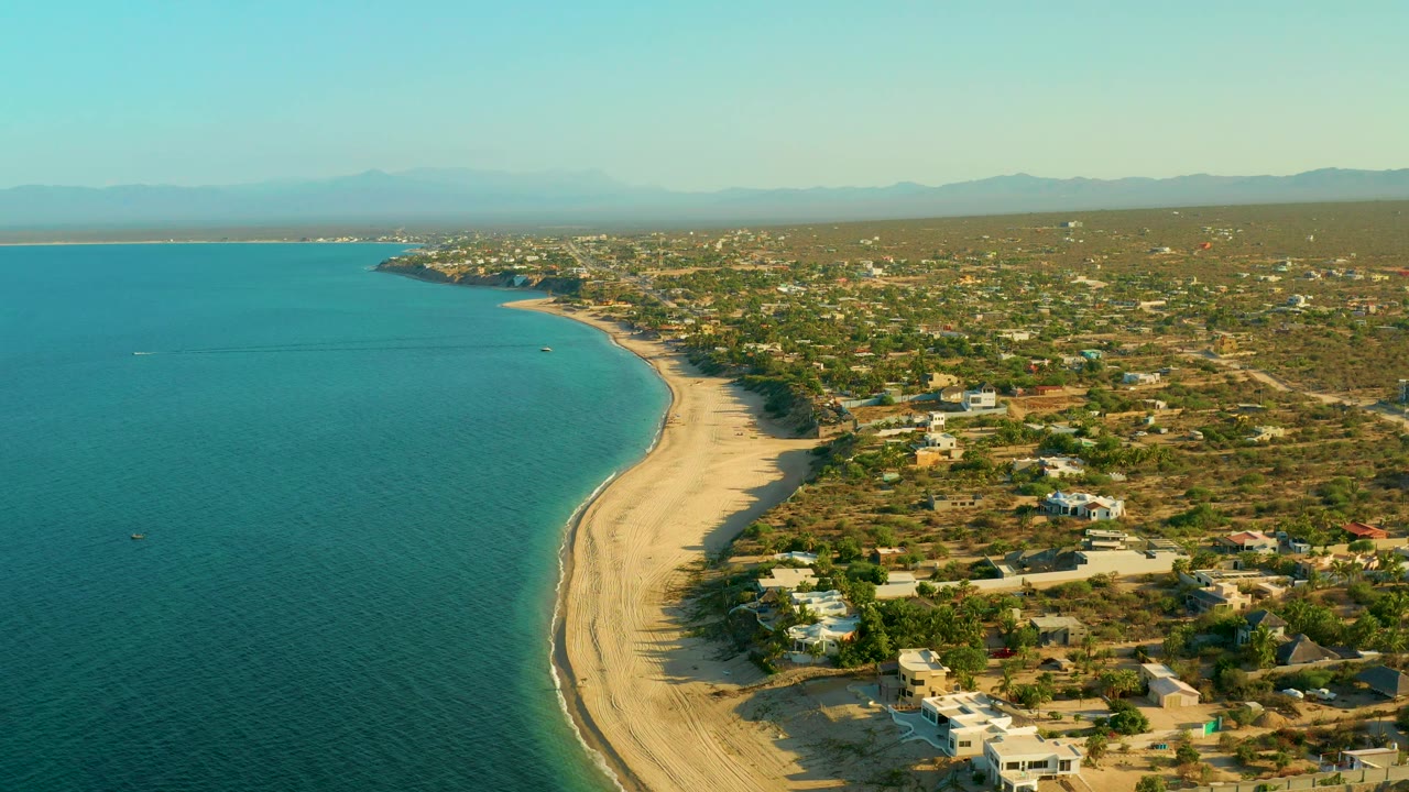 Wide aerial frame over El Sargento La Ventana, Mexico, with the ocean filling the left side, the beach running down the center, desert lots and buildings to the right, and no visible people.