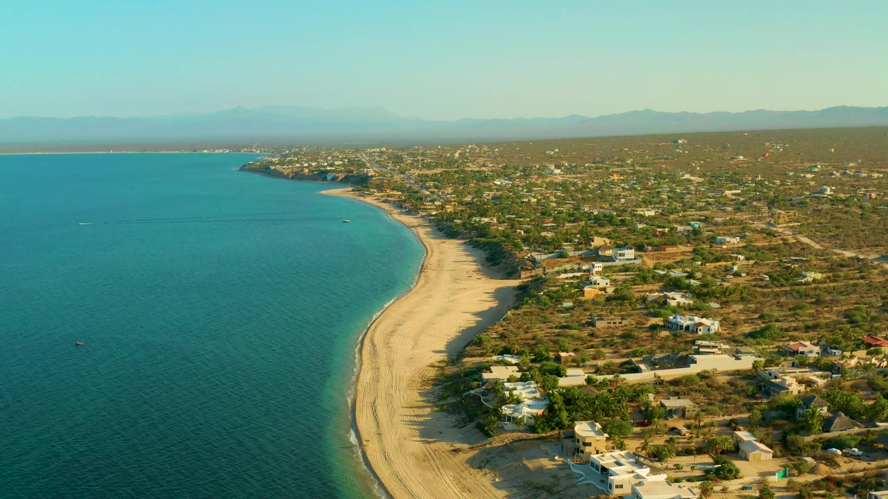 Aerial shoreline view with calm sea, pale beach, homes, and inland desert