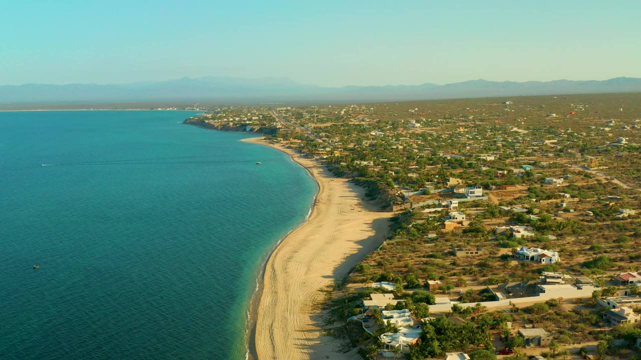 High aerial view over El Sargento La Ventana, Mexico, with turquoise ocean on the left, a curving sand beach in the center, scattered coastal homes on dry terrain to the right, hazy mountains beyond, and no visible people.
