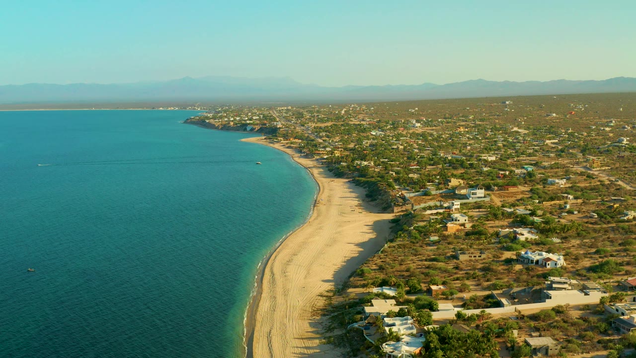 Wide aerial view in El Sargento La Ventana, Mexico, with calm turquoise sea to the left, a long sandy shoreline curving through the frame, white buildings among scrubland to the right, distant mountains, and no visible people.