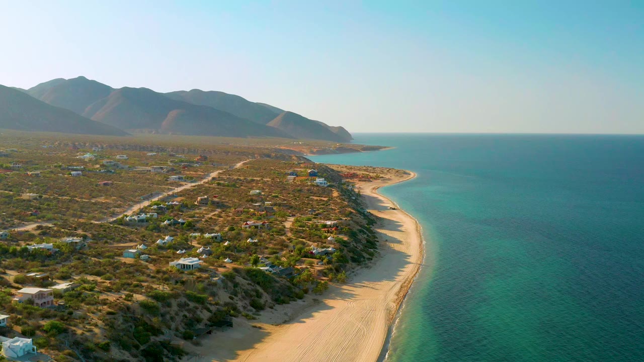 A wide aerial frame over El Sargento La Ventana, Mexico shows a curving sandy beach beside blue ocean, scrubland with scattered homes, distant mountains, and no visible people.
