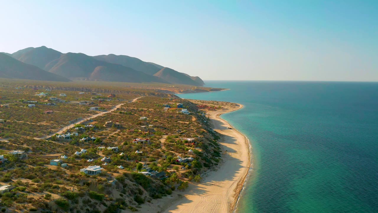 Aerial frame over El Sargento La Ventana, Mexico, showing a curved sand beach, calm blue water, sparse residential structures, dirt tracks, mountain ridges, and no visible people.