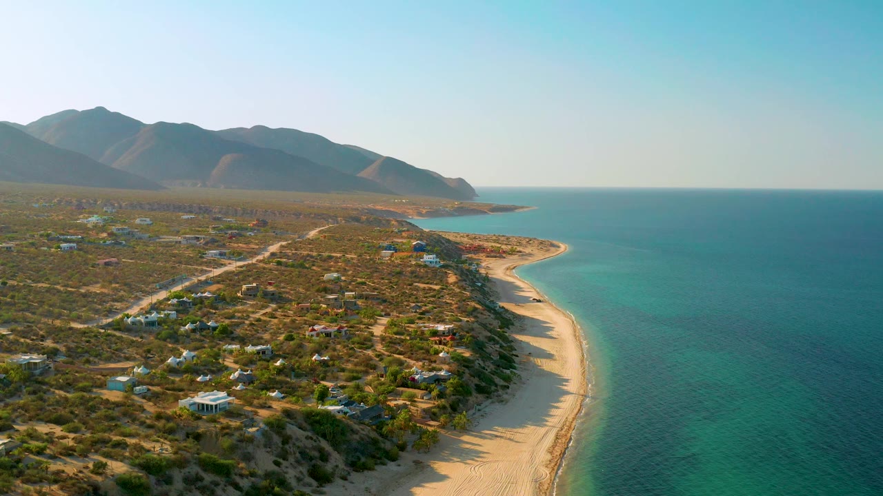 Wide aerial view of El Sargento La Ventana, Mexico, with the ocean filling the right side, beach and rocky shore through the center, low homes in scrubland, mountains behind, and no visible people.
