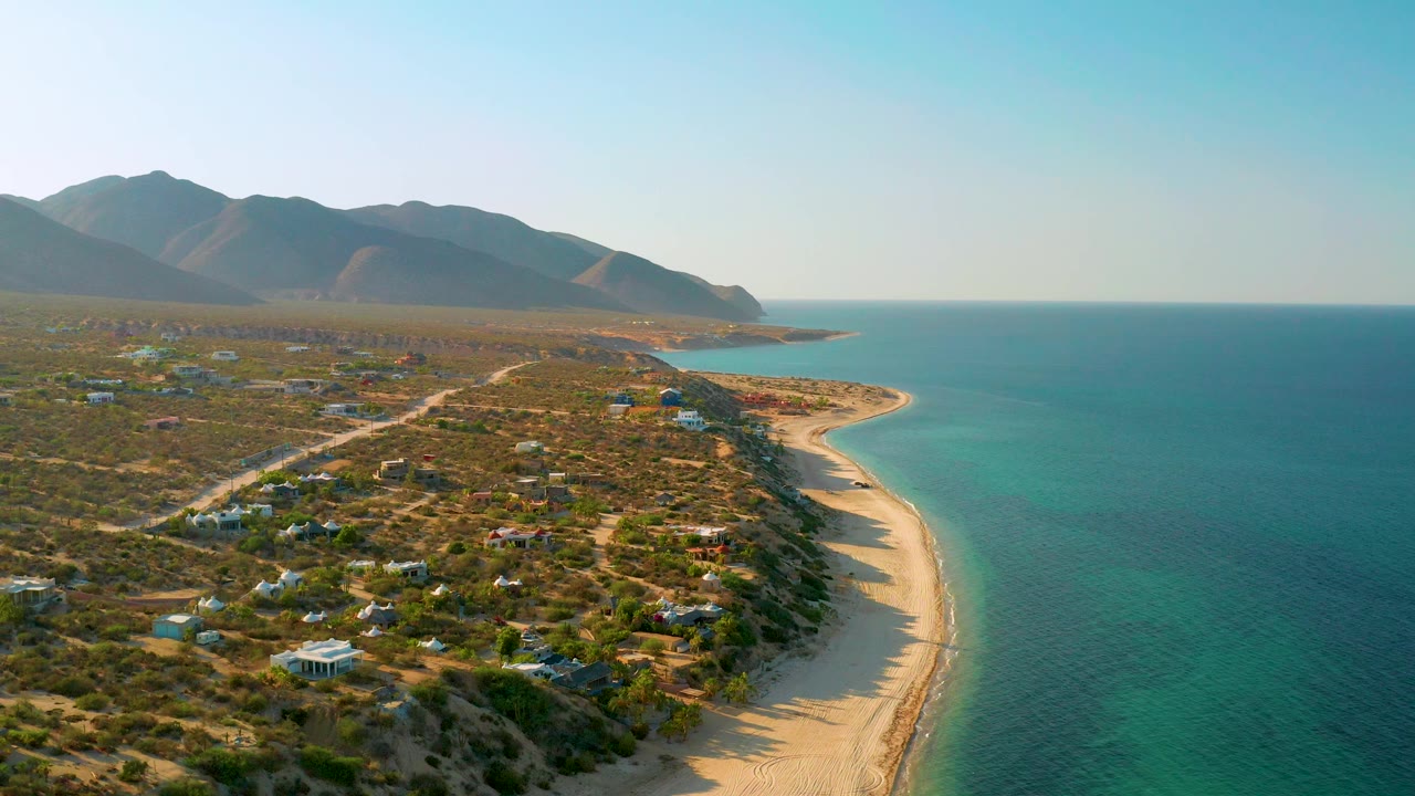 High coastal view with beach, turquoise shallows, desert vegetation, homes, and mountains.