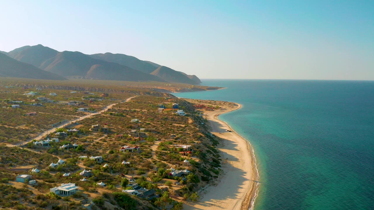 Aerial view over El Sargento La Ventana, Mexico, with turquoise sea on the right, a pale curved beach below, scattered coastal buildings on dry scrubland, distant mountains, and no visible people.