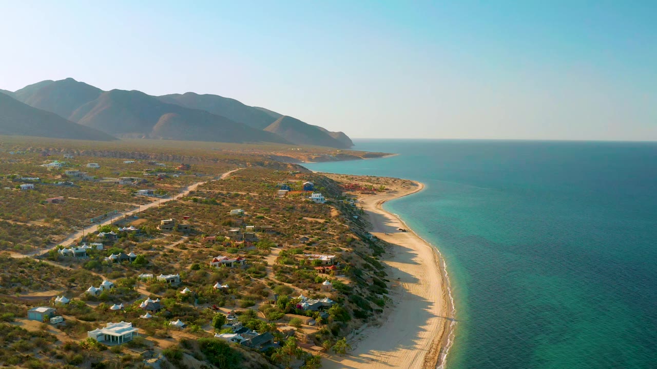 High aerial frame from El Sargento La Ventana, Mexico, showing the shoreline running diagonally past small buildings, dirt roads, dry vegetation, blue-green water, distant mountains, and no visible people.