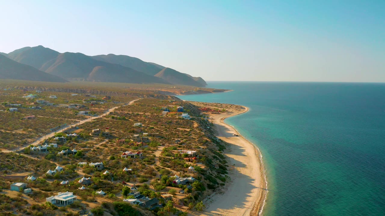 Aerial view in El Sargento La Ventana, Mexico, with shallow turquoise water meeting a narrow sand beach, low scrubland and scattered structures inland, a mountain ridge across the background, and no visible people.