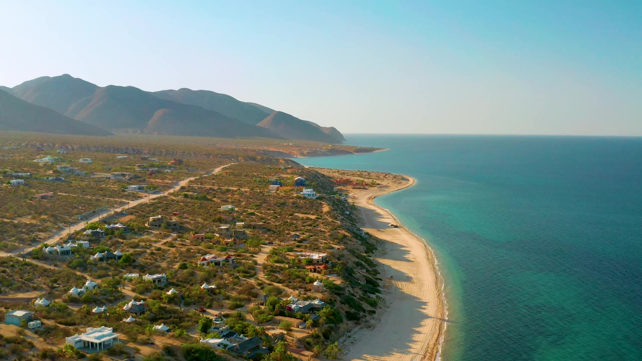 Aerial coastline with dry scrubland, beach, sea, and hazy mountains.