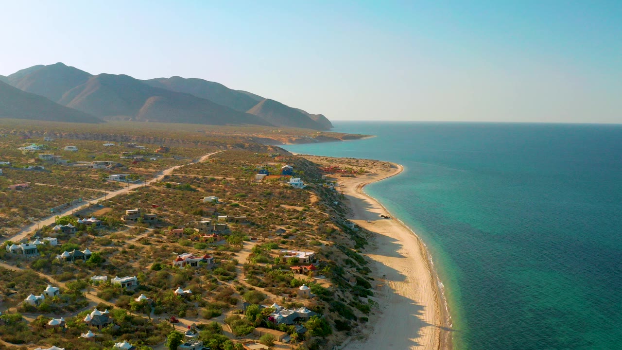 Opening aerial frame over El Sargento La Ventana, Mexico, with scattered homes and dirt roads across scrubland, a sandy beach at right, blue water beyond it, distant mountains, and no visible people.
