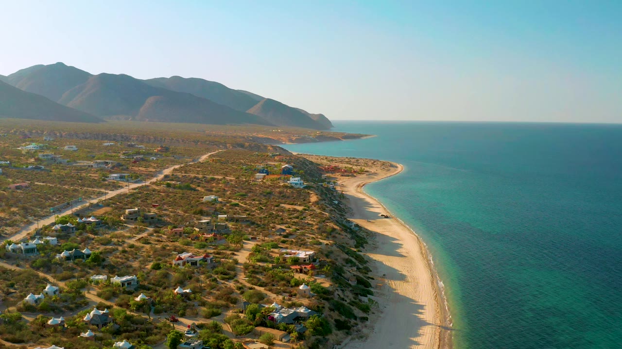 High coastal view with curved beach, blue sea, brushy land, small buildings, and mountains.