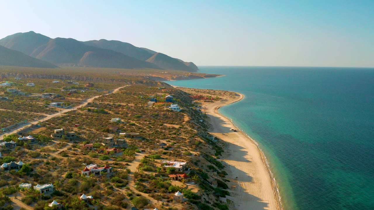 Opening aerial view over El Sargento La Ventana, Mexico, with scattered homes and dirt roads on dry coastal terrain, a pale beach curving beside blue ocean, mountains in back, and no visible people.