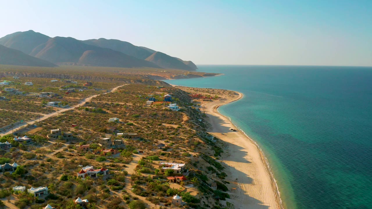 Drone view over El Sargento La Ventana with the empty sandy beach running down the right side, teal water beside it, sparse homes and roads on scrubland to the left, and no visible people.