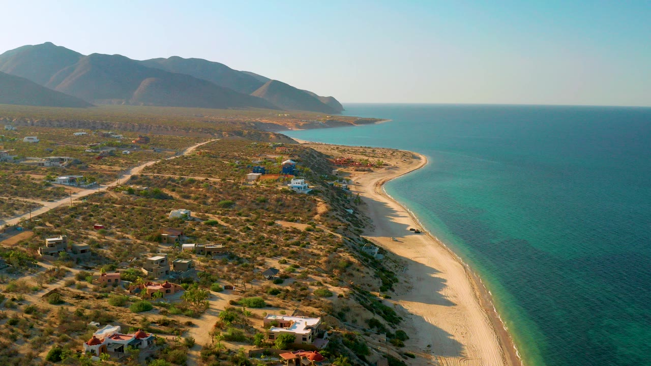 Opening aerial frame over El Sargento La Ventana, Mexico, showing scattered desert homes, a curved sandy beach, turquoise sea, and dark mountains, with no visible people.