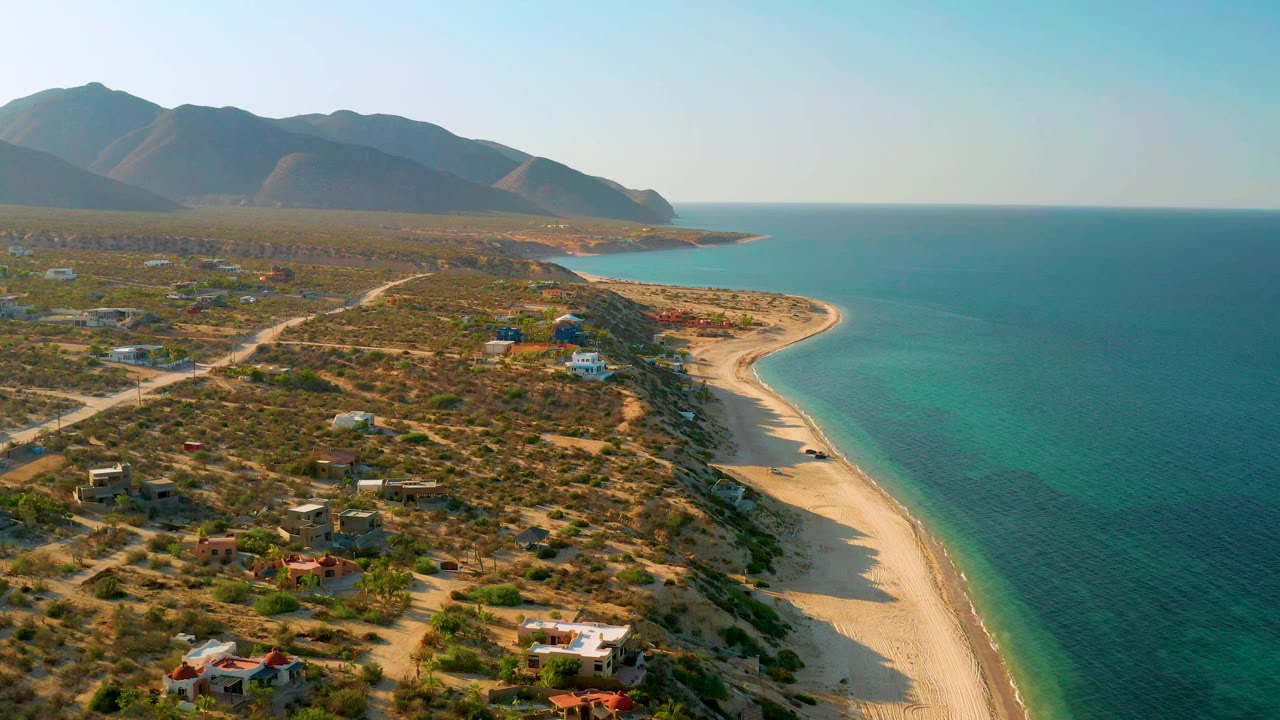 Aerial frame over El Sargento La Ventana, Mexico, with the shoreline sweeping toward the mountains while dirt roads and small homes sit across the dry foreground, with no visible people.