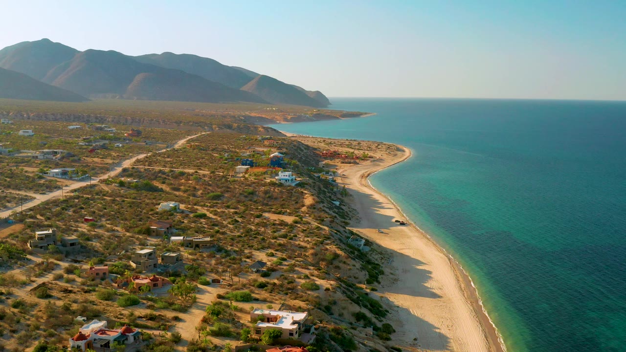 Aerial frame in El Sargento La Ventana, Mexico, showing more open sea to the right, a pale beach edge, scrubland lots, and distant mountains under warm light, with no visible people.