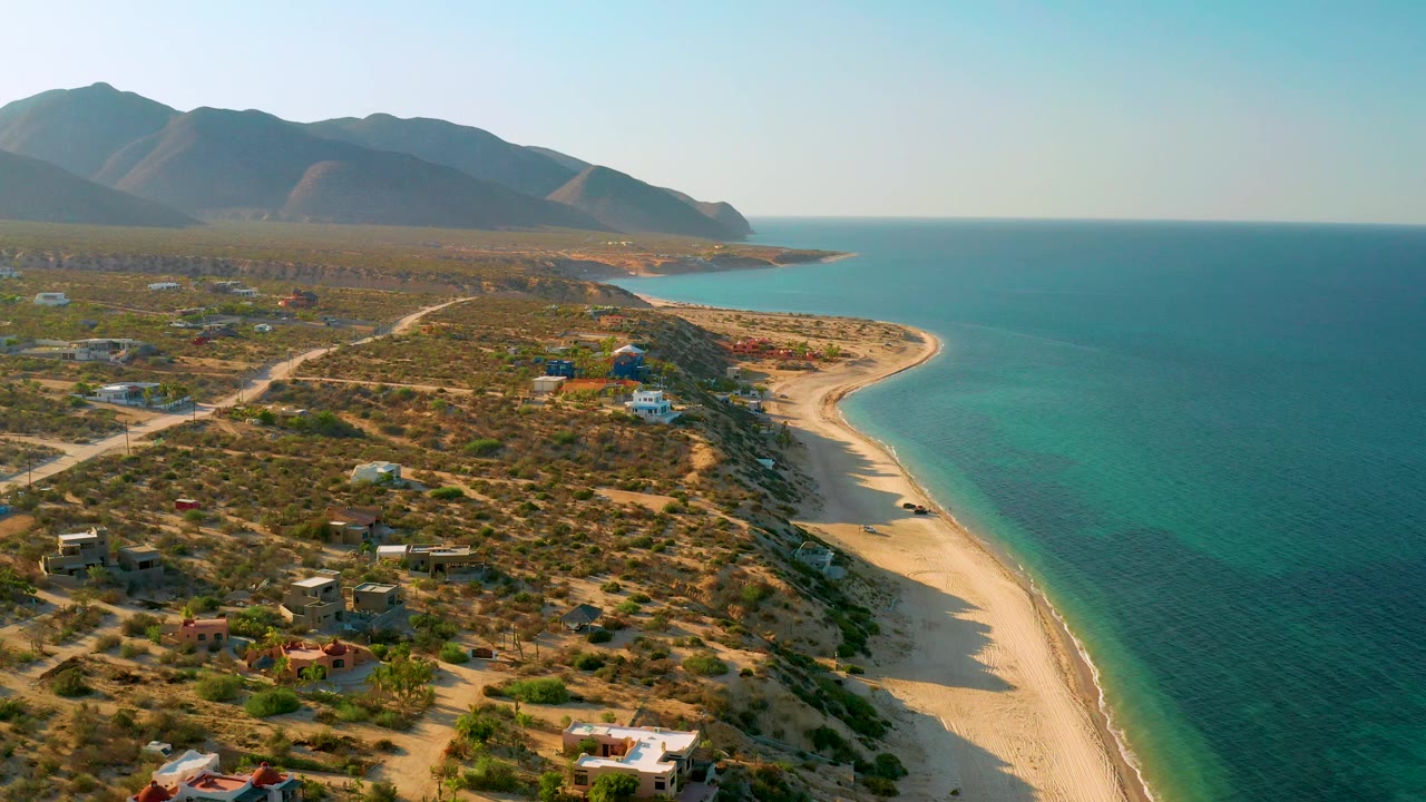 Wide aerial coastline with desert neighborhood, curving beach, turquoise sea, and mountains.