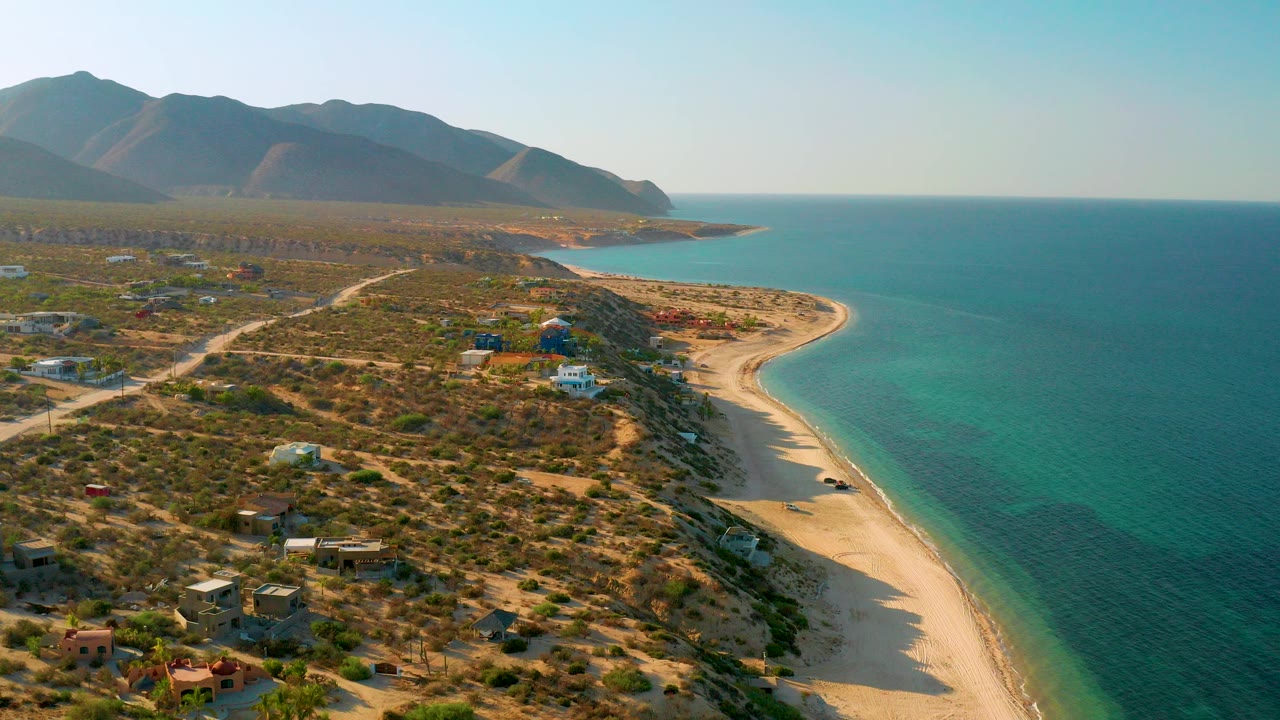 A high aerial frame over El Sargento La Ventana, Mexico shows a curved golden beach, turquoise green sea, scattered low buildings, dirt roads, mountains, and no clearly visible people.