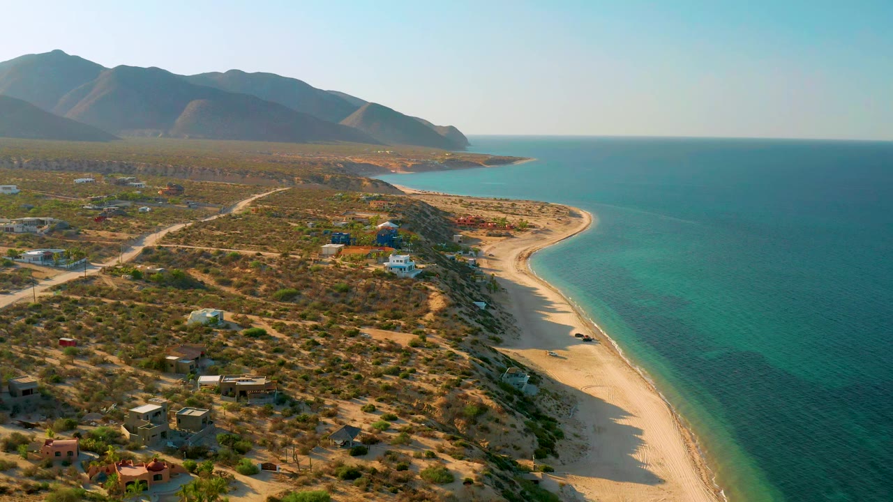 Drone view of shoreline, turquoise sea, desert brush, homes, and mountains.