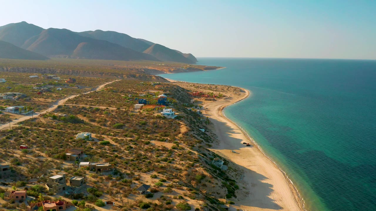 A wide aerial frame shows the beach arc running toward the distance, clear green-blue water on the right, low desert growth and roads on the left, mountain ridges behind, and no clearly visible people.